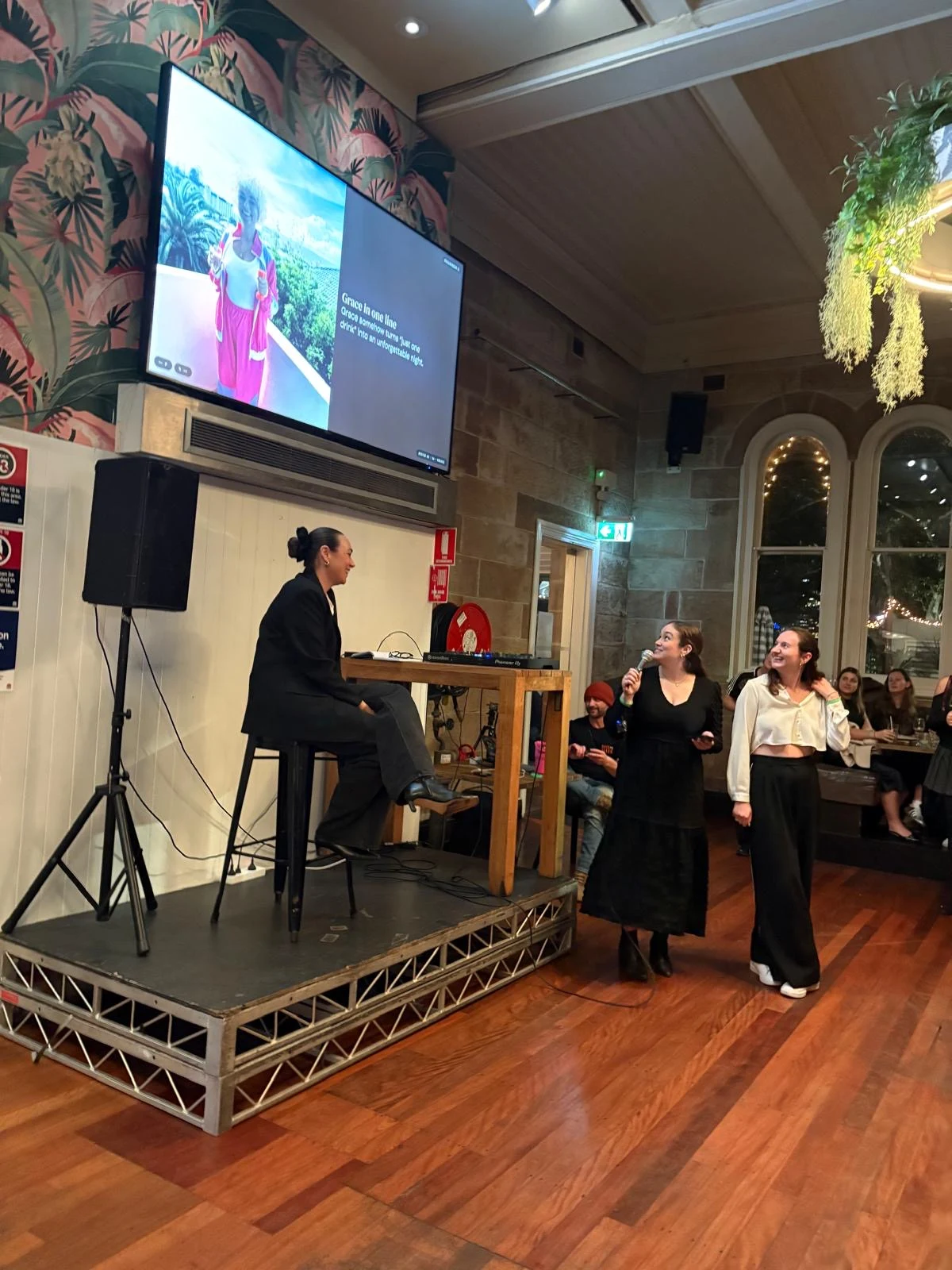 A mate at the mic pitching her single friend at The Greenwood, her slide on the screen behind